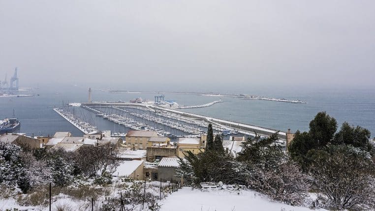 Vue panoramique du port de Sète sous la neige, quais, immeubles colorés et bateaux figés au pied des collines sous un ciel gris d’hiver.