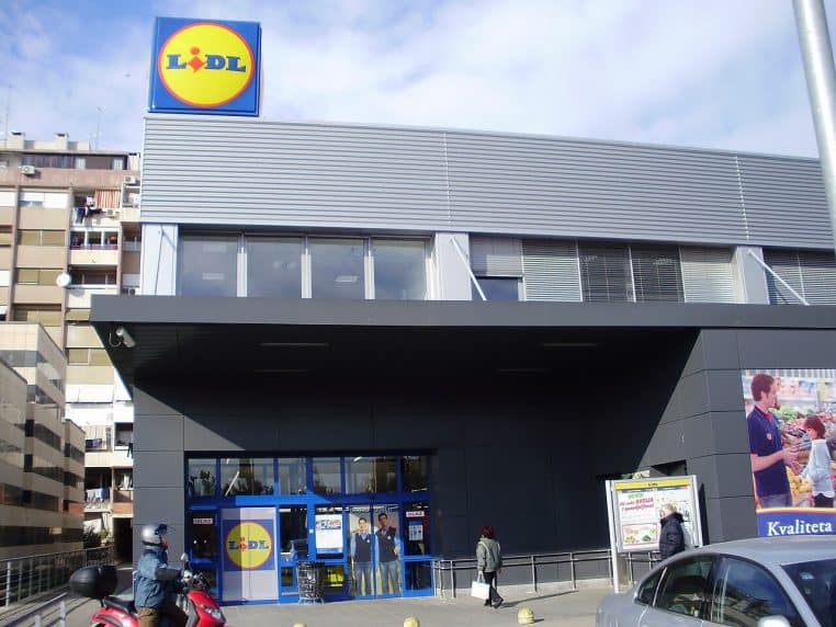 Entrée d’une enseigne Lidl avec portes vitrées et logo, prise en extérieur par temps clair, photo horizontale.