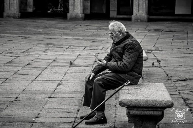 Un homme âgé assis sur un banc consulte son smartphone dans la rue, illustrant la maîtrise des outils numériques par certains seniors.