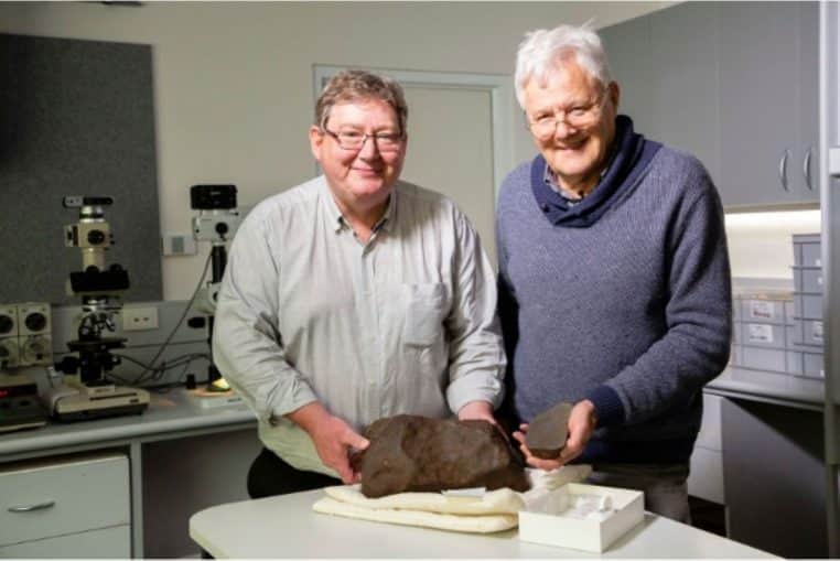 Deux géologues du Museums Victoria posent dans un laboratoire derrière un gros fragment sombre de la météorite de Maryborough posé sur une table blanche.