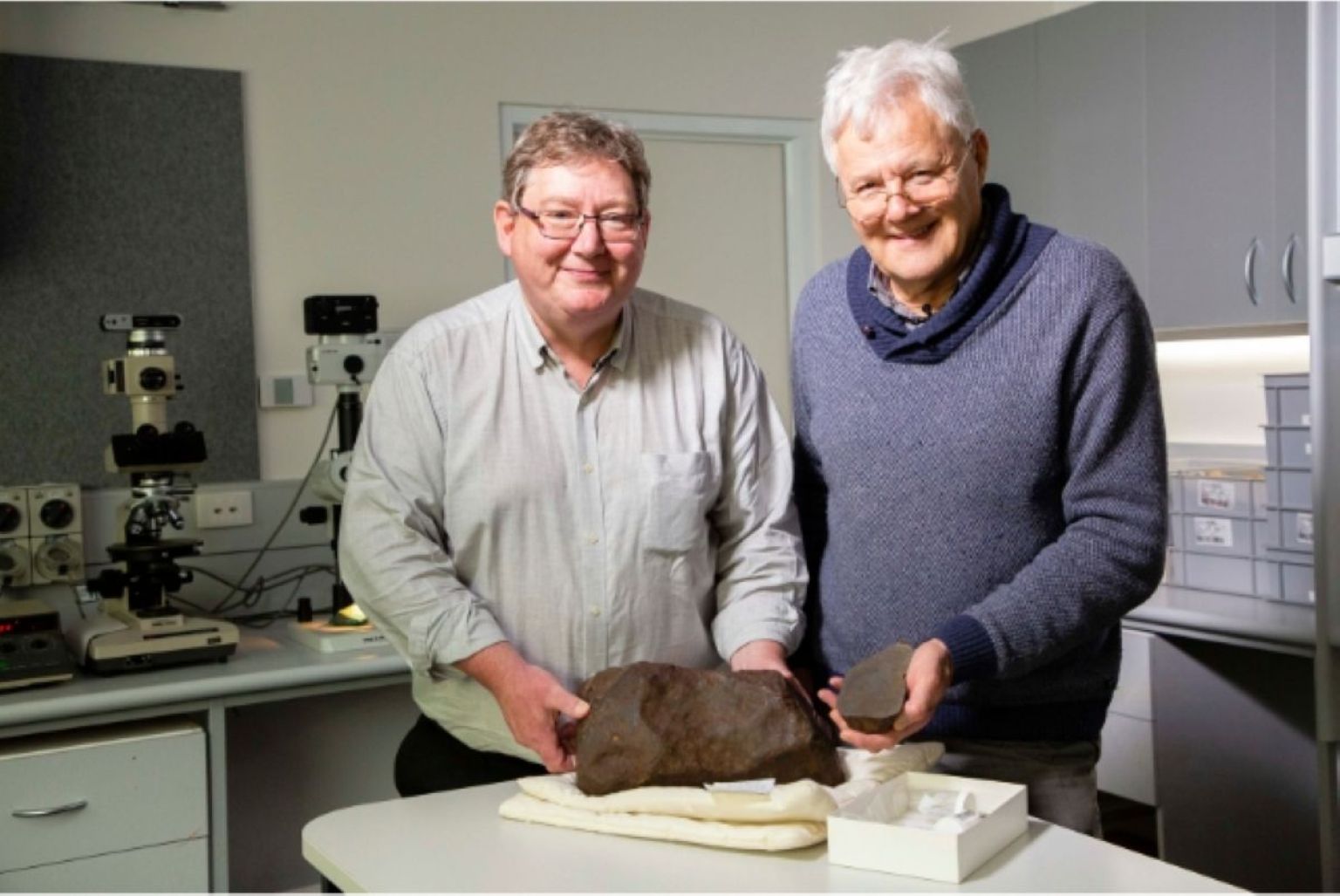 Deux géologues du Museums Victoria posent dans un laboratoire derrière un gros fragment sombre de la météorite de Maryborough posé sur une table blanche.
