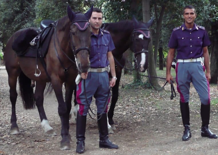 Deux agents de police montés posent avec leurs chevaux dans un parc, tenue sombre, chemin boisé en arrière-plan