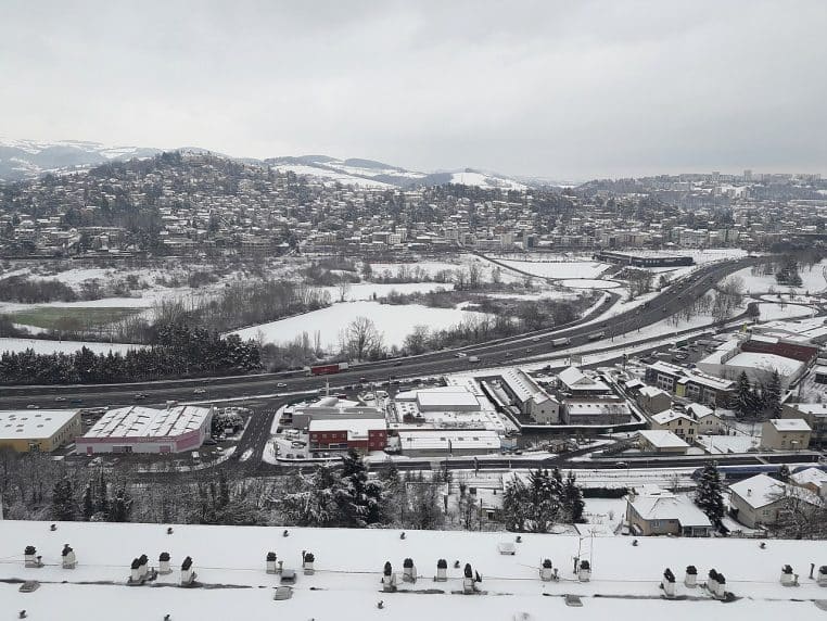 Vue panoramique de Saint-Étienne sous la neige, autoroute visible et toits blanchis dans une lumière grise d’hiver.