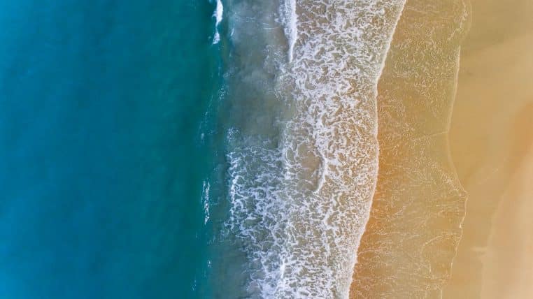 Image drone verticale d’une plage où la mer bleue profonde se heurte à un rivage doré, avec l’écume blanche dessinant une frontière nette.