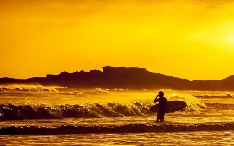 Silhouette d’un surfeur au coucher du soleil, planche sous le bras, reflets dorés sur les vagues et horizon sombre