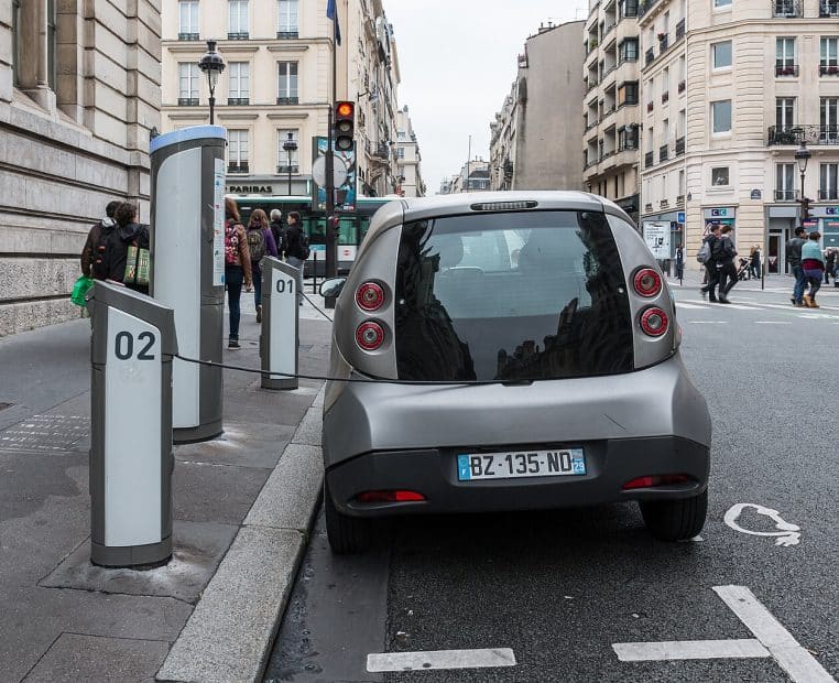 Voiture électrique branchée sur une borne de recharge à Paris, photographiée en plan large avec la rue en arrière-plan.