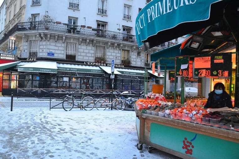 Marché de quartier à Paris sous la neige, trottoir blanchi et traces de pas devant des immeubles haussmanniens.