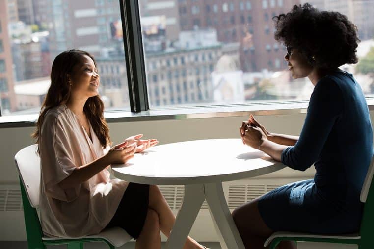 Deux femmes discutent face à face dans un café lumineux avec vue sur la ville, concentrées l’une sur l’autre dans un échange chaleureux.