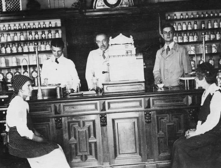 Comptoir d’une pharmacie ancienne avec étagères de flacons, scène de vente et de préparation au centre