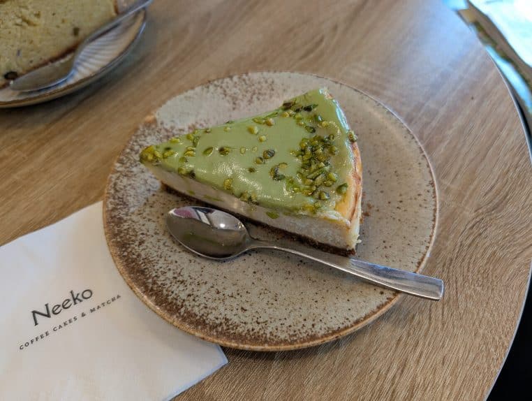 Part de cheesecake à la pistache surmontée d’éclats verts, posée sur une assiette blanche, évoquant la crème pistache de la bûche.