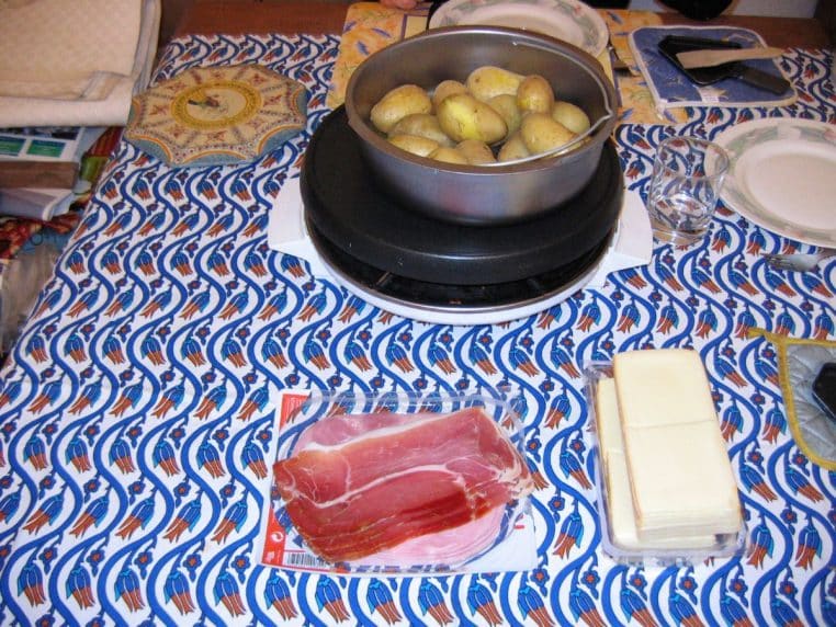 Appareil à raclette sur une table, avec pommes de terre, tranches de fromage et charcuterie prêtes à servir.