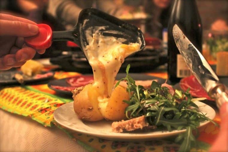 Fromage à raclette fondu versé sur des pommes de terre, avec salade verte sur le côté, pendant un repas partagé.
