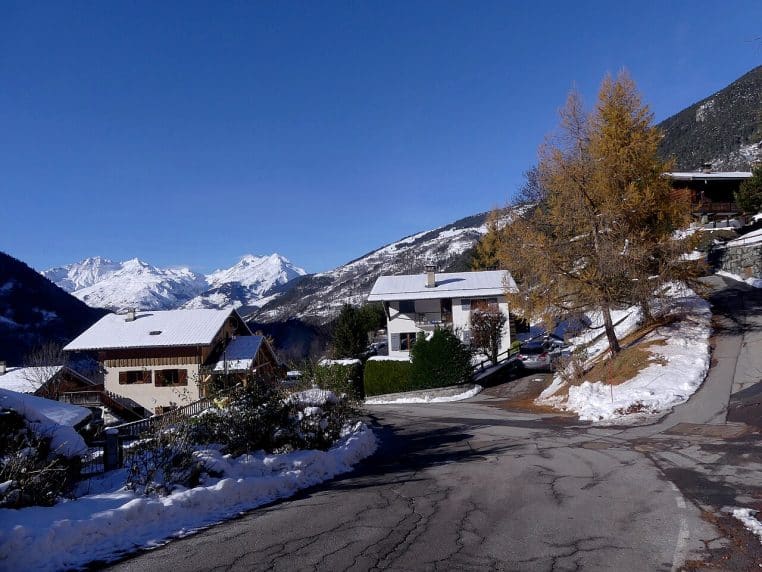 Route en Savoie après chutes de neige, virage en pente avec maisons et montagnes au fond sous ciel bleu.