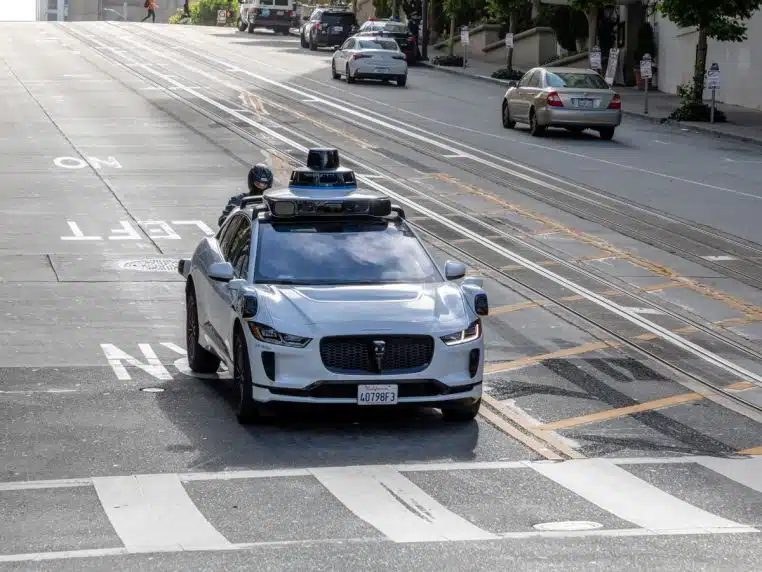 Voiture autonome Waymo blanche photographiée sur California Street à San Francisco, au milieu d’un trafic urbain typique avec immeubles et câbles aériens.