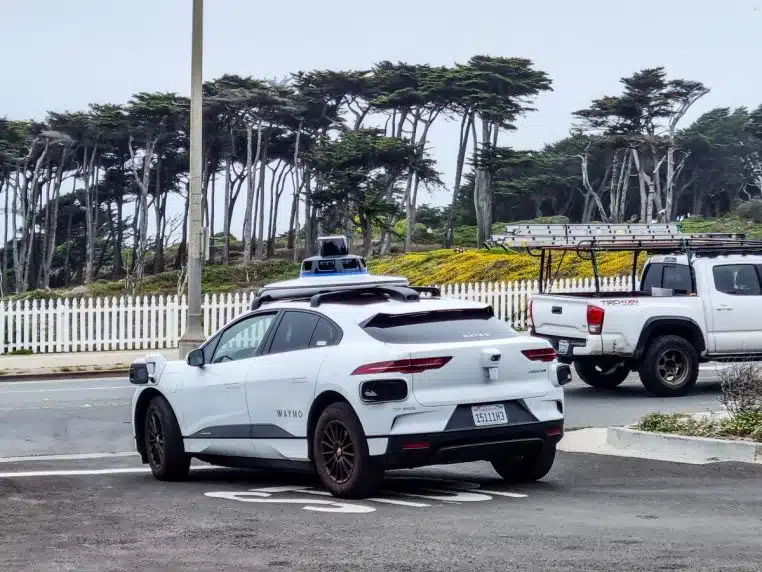Voiture autonome Waymo stationnée sur un parking de San Francisco, vue de côté, avec océan et collines en arrière-plan par temps dégagé.