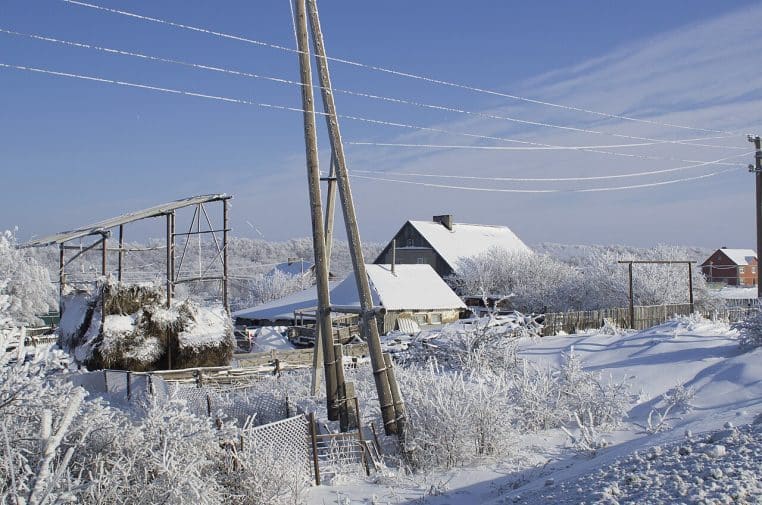 Maisons de campagne et clôtures recouvertes de neige sous un ciel bleu, avec givre visible et reliefs au fond.
