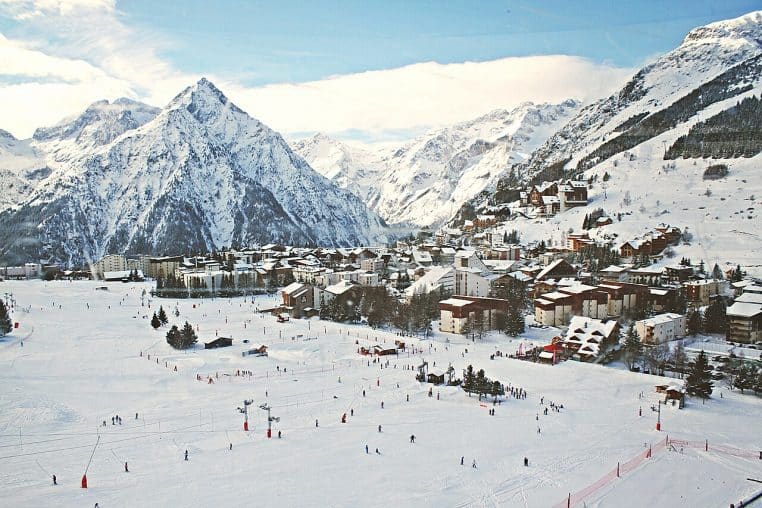Station de montagne et village enneigés au pied de sommets, sous un ciel lumineux, panorama large et hivernal.
