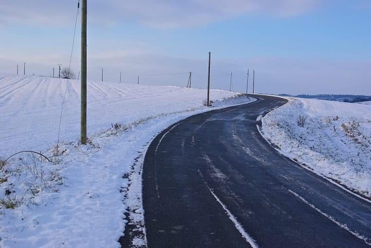 Route de campagne bordée de neige en fin de journée, ciel clair et chaussée sombre dans un décor hivernal.