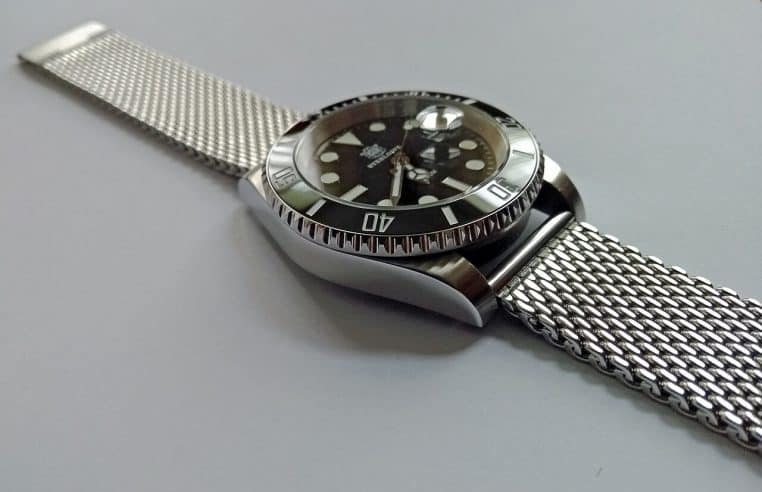 Montre sportive en acier vue de profil sur bracelet maille, posée sur fond gris, verre et lunette visibles.