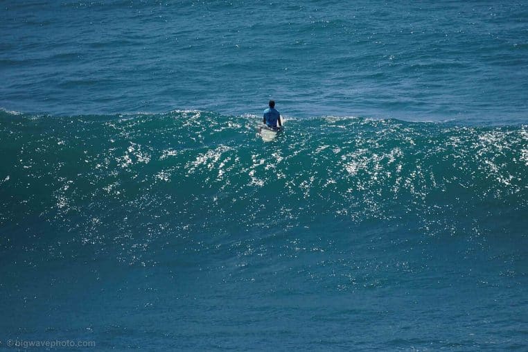 : Surfeur assis sur sa planche au large, vu de loin, une vague luit au soleil et l’océan bleu remplit le cadre
