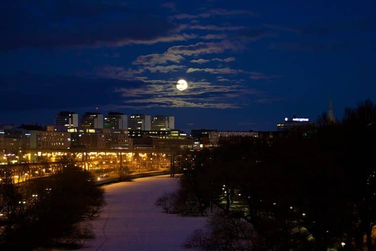 Vue nocturne d’une ville enneigée éclairée par les lumières urbaines, avec une pleine Lune brillante se levant derrière les nuages bas dans un ciel bleu profond.