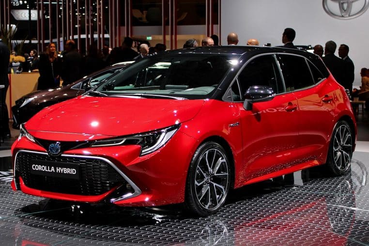 Toyota Corolla Hybrid rouge exposée au Mondial de l’Auto, vue de trois quarts avant, entourée de visiteurs et de stands lumineux.