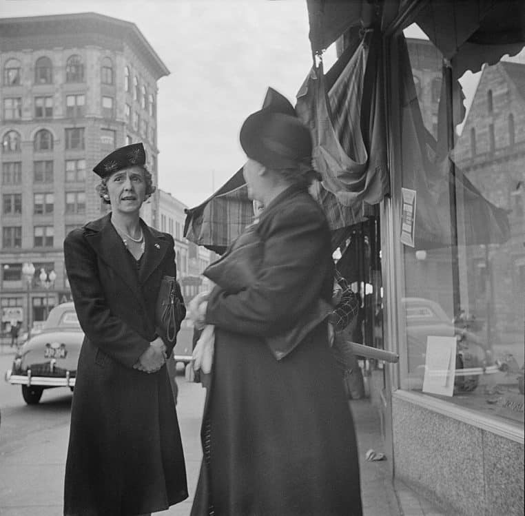 Deux femmes debout sur un trottoir en 1941 discutent face à face, manteaux d’époque, plongées dans une conversation animée.