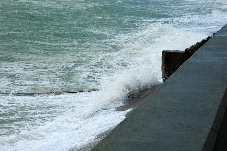 Vague frappant la digue à Veulettes-sur-Mer, embruns visibles et mer agitée sous un ciel gris