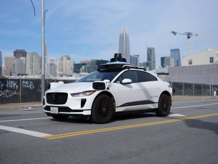 Voiture autonome Waymo blanche roulant sur une rue en pente bordée de maisons colorées à San Francisco, observée depuis l’arrière dans la circulation urbaine.