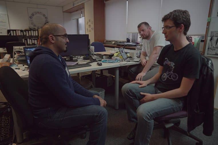 Réunion informelle dans un bureau, trois personnes assises face à face près d’ordinateurs et dossiers sur une table.
