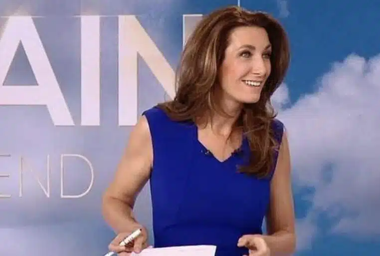 Anne-Claire Coudray souriante en plateau TV, robe bleue, tenant des feuilles, fond de décor ciel et lettrage.
