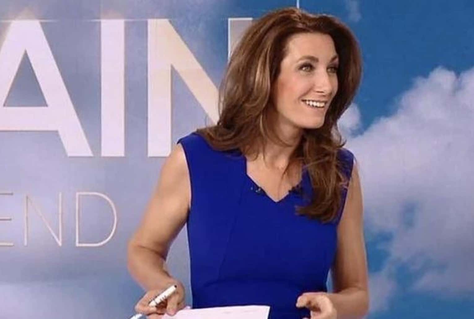 Anne-Claire Coudray souriante en plateau TV, robe bleue, tenant des feuilles, fond de décor ciel et lettrage.
