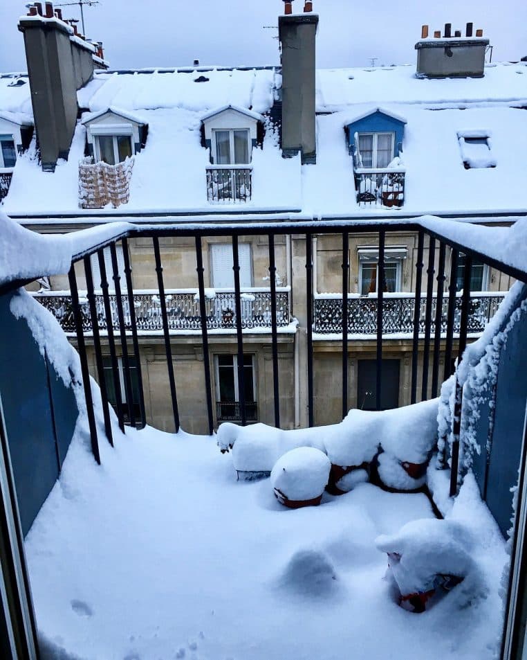 Balcon parisien recouvert d’une épaisse couche de neige, vue sur les toits haussmanniens et cheminées sous un ciel gris uniforme