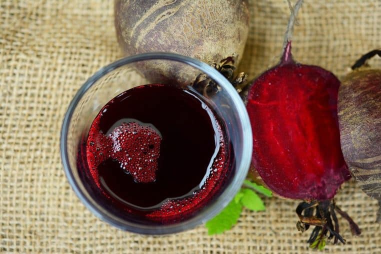 Verre de jus de betterave rouge vu de dessus sur une toile de jute, entouré de betteraves crues entières et coupées, prêtes à être consommées.