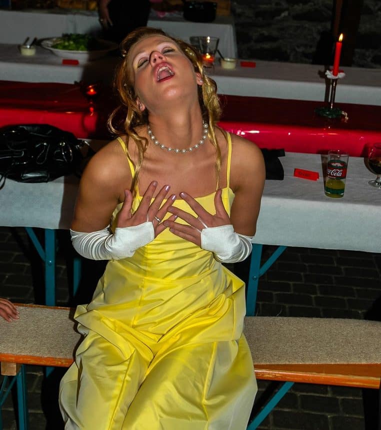 Une femme en robe jaune de soirée, assise à une table de fête, penche la tête en arrière en posant les mains sur sa poitrine