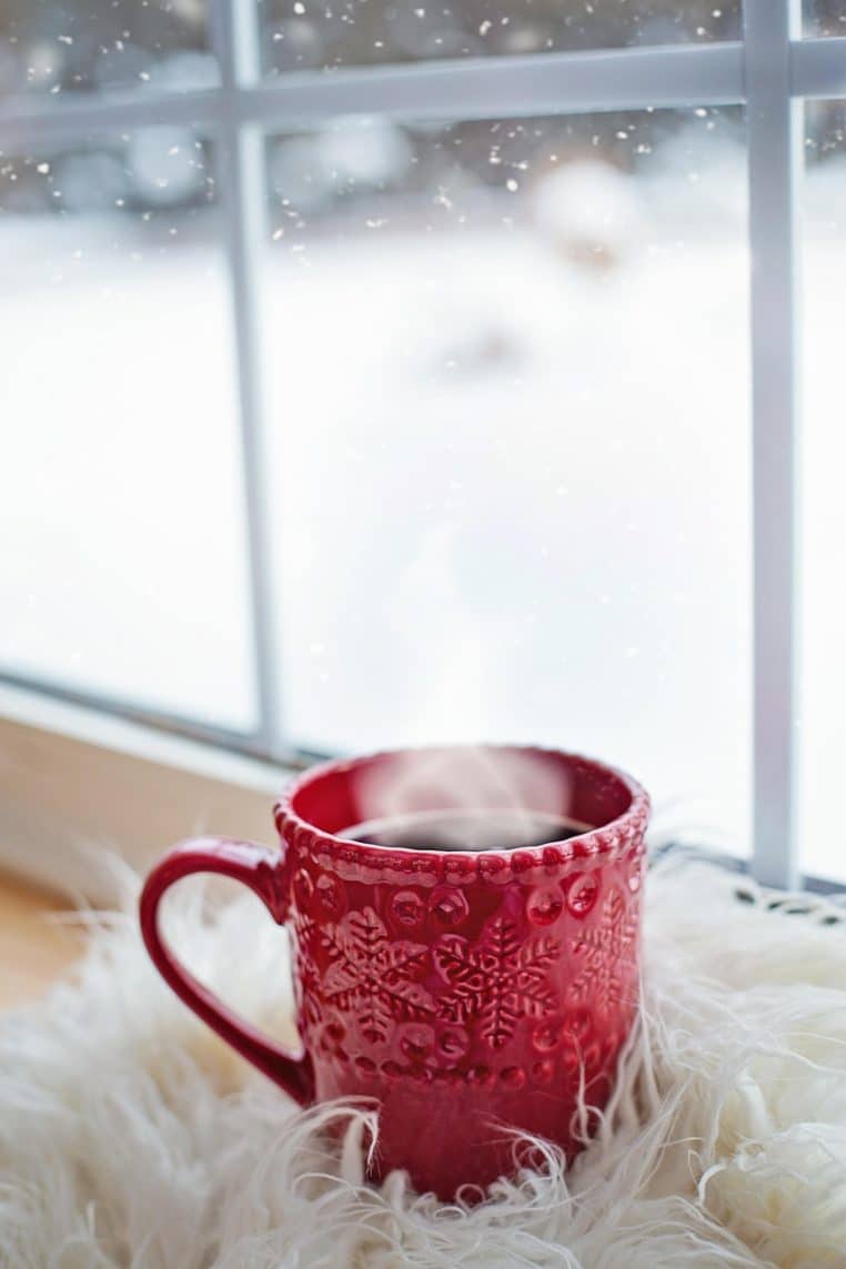 Tasse de café fumant posée sur un rebord de fenêtre, neige abondante visible à travers la vitre d’un salon en hiver