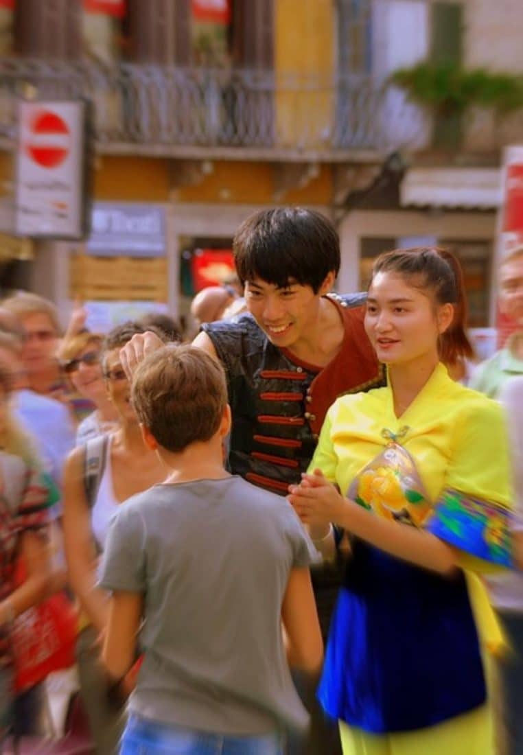 Un jeune garçon s’avance vers deux artistes souriants en costume coloré, au milieu d’une foule floue dans une rue animée