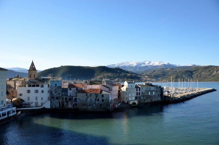 Village portuaire méditerranéen aux façades pastel bordant une mer calme, avec montagnes enneigées en arrière-plan sous un ciel bleu limpide.