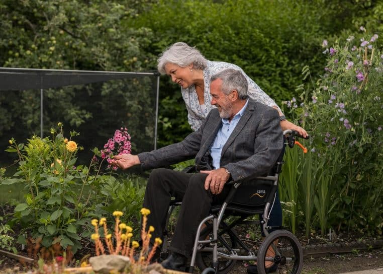 Couple de seniors dans un jardin fleuri, l’homme assis dans un fauteuil roulant manipulant des plantes avec l’aide de sa compagne souriante.