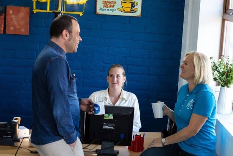 Trois collègues échangent en souriant autour d’un bureau d’ordinateur, tasses à la main, dans un bureau moderne aux murs bleus