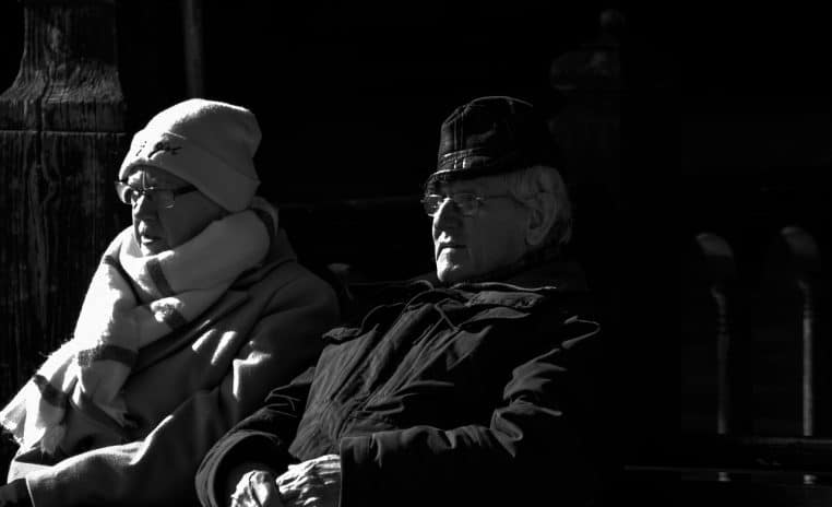 Couple de seniors assis côte à côte sur un banc en plein air, tenant leurs mains, symbole de solidarité face aux changements de pension