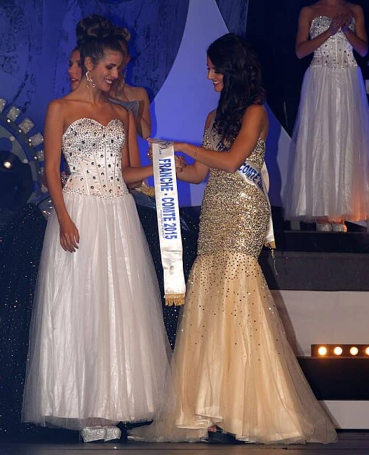 Deux candidates en robe longue échangent l’écharpe Miss Franche-Comté 2015 sur scène, sous les projecteurs d’un concours régional de beauté.