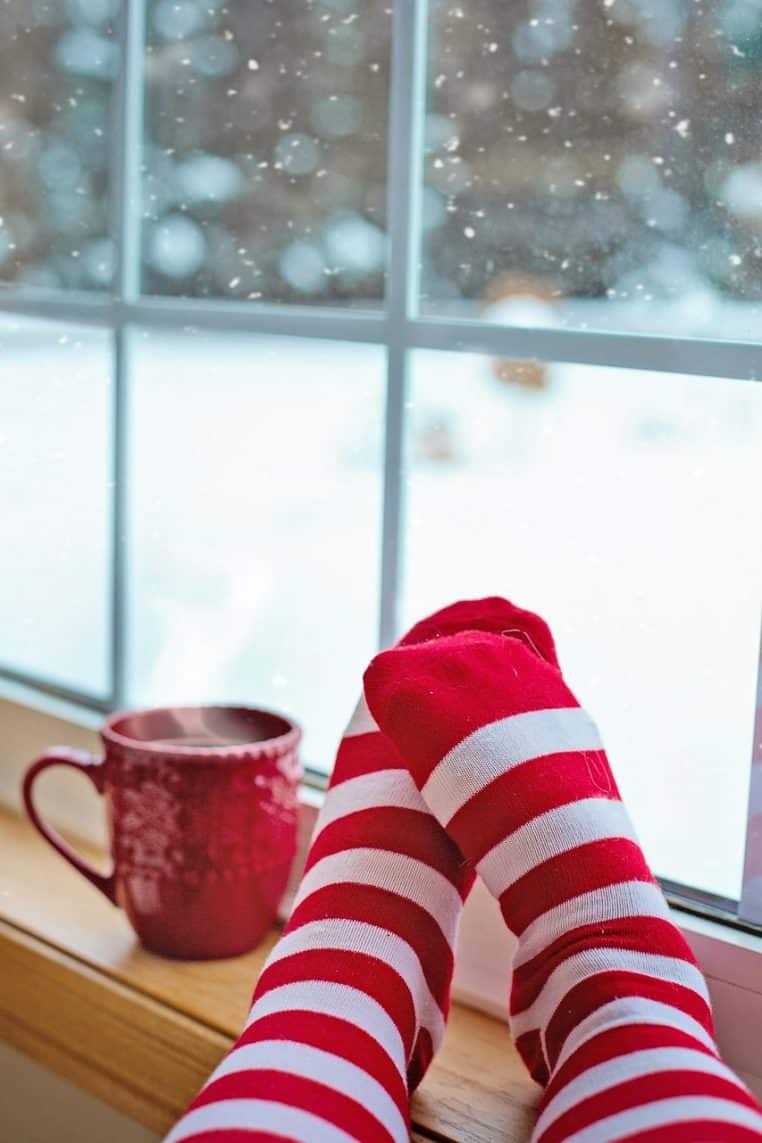 Pieds emmitouflés dans des chaussettes près d’un mug de café, grande fenêtre donnant sur un paysage enneigé en hiver