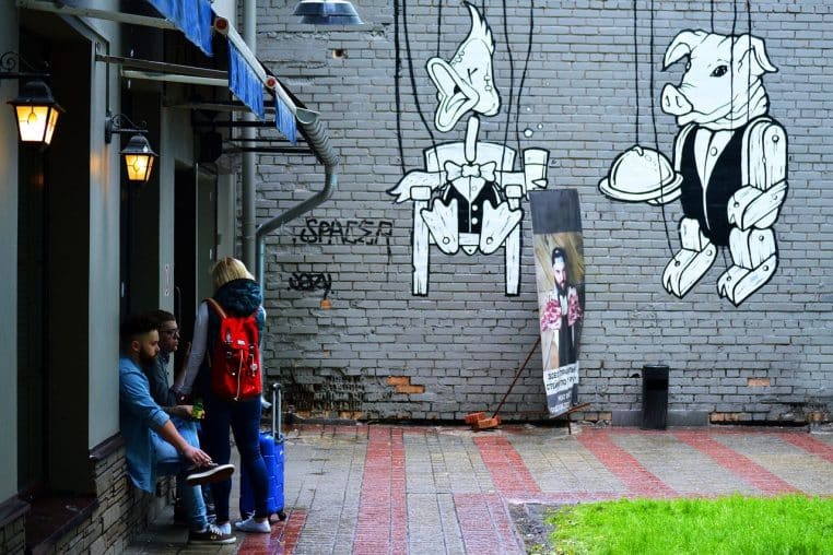 Trois amis discutent devant un mur de briques couvert de graffitis noirs, dans une rue humide, l’un assis et deux debout