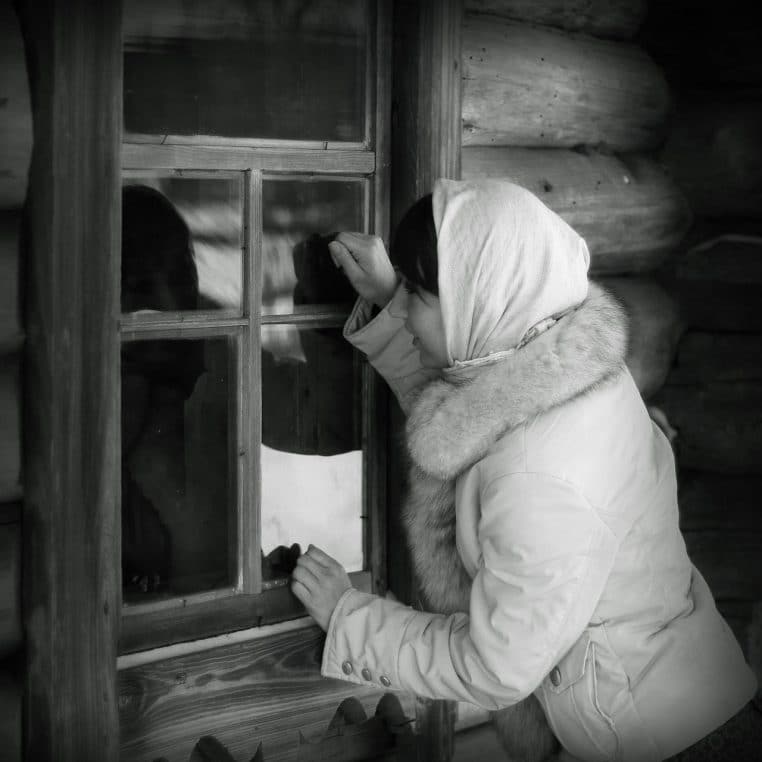 Jeune femme en pull regardant par une fenêtre en hiver, givre sur la vitre et paysage enneigé visible à l’extérieur du logement