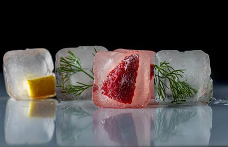 Gros plans sur des glaçons transparents renfermant des morceaux de fruits et des herbes, alignés sur une surface brillante