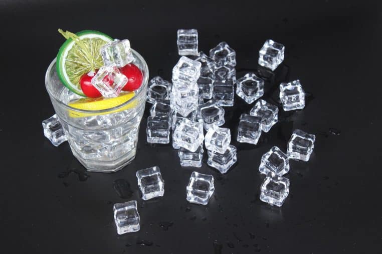 Verre rempli de glaçons avec tranches de citron vert et cerises, entouré de nombreux cubes de glace sur une surface sombre