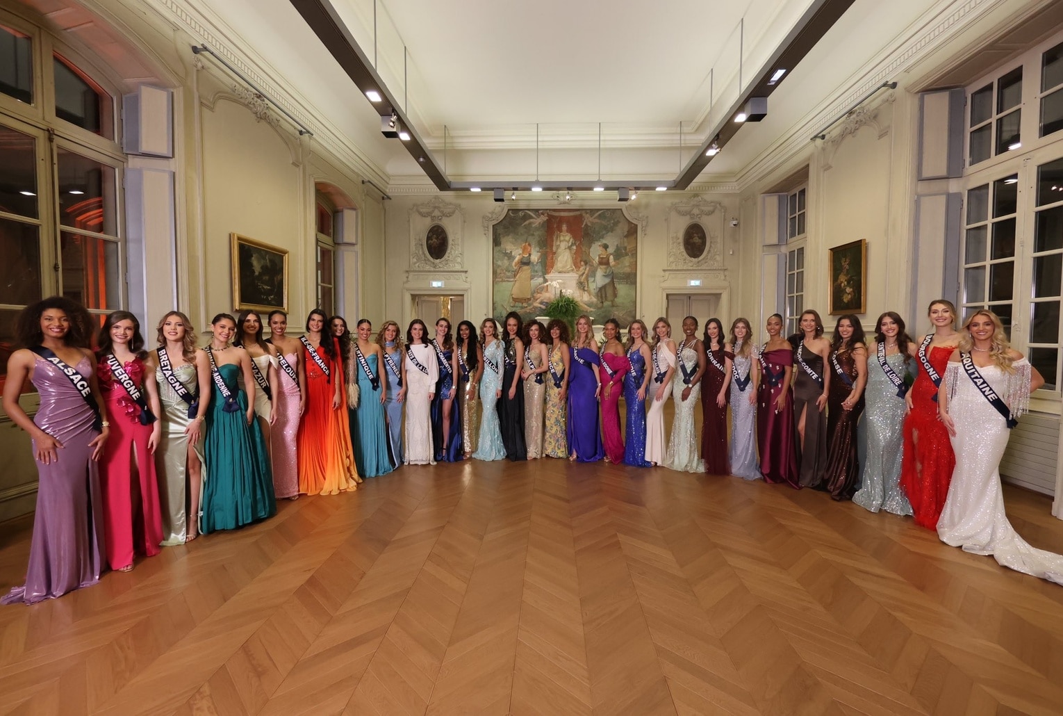 Les trente candidates à Miss France 2026 posent côte à côte en robes de soirée colorées dans un salon au parquet en chevron.