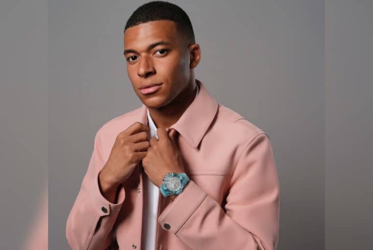Portrait de Kylian Mbappé en veste rose, montrant au poignet une montre bleu translucide à cadran squeletté.