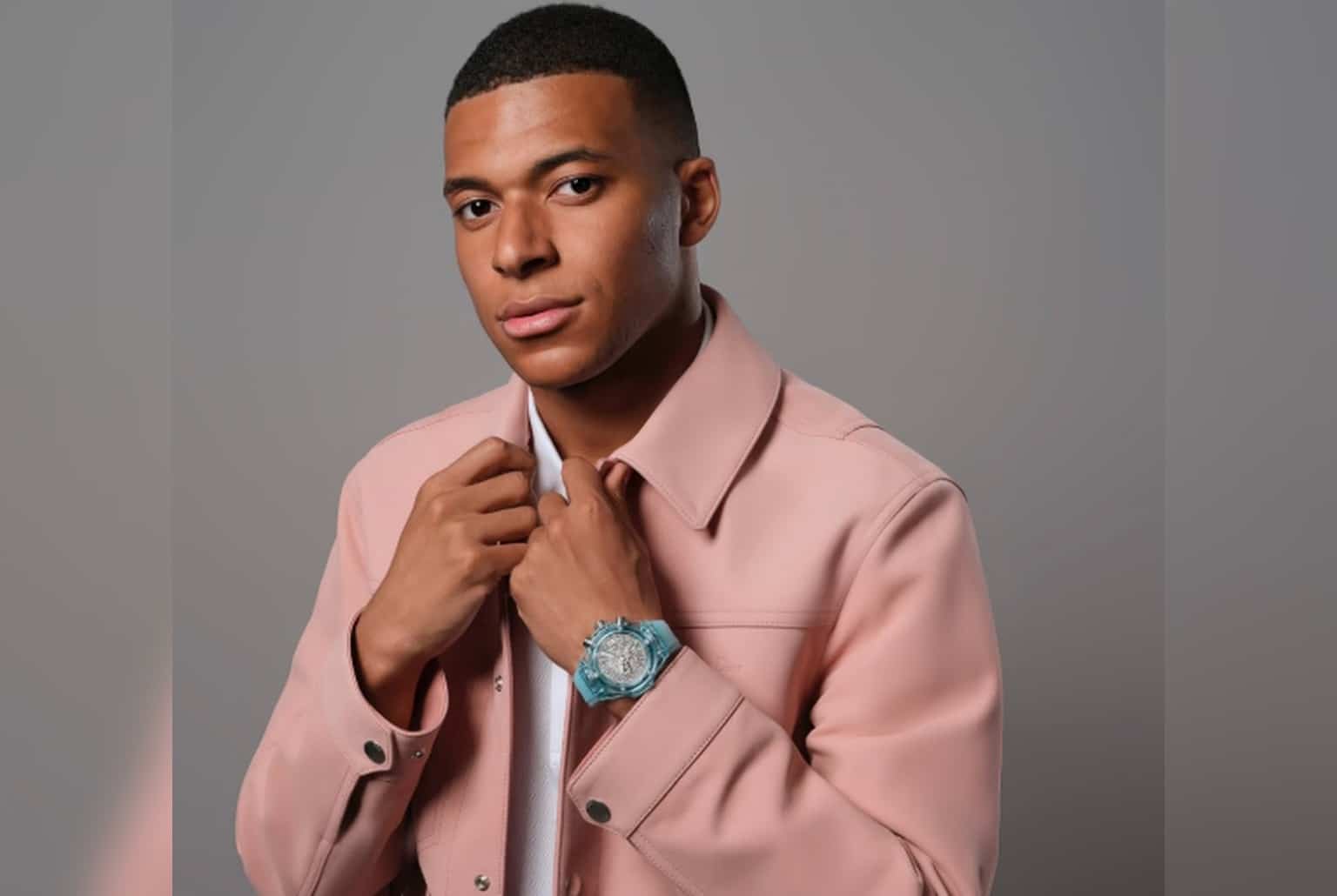 Portrait de Kylian Mbappé en veste rose, montrant au poignet une montre bleu translucide à cadran squeletté.